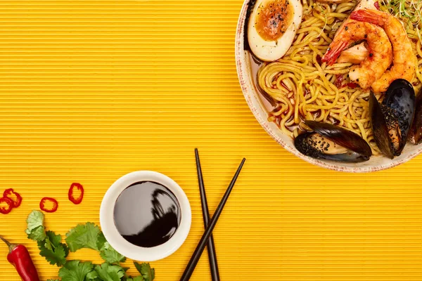 Vue du dessus des ramen de fruits de mer près des ingrédients frais, de la sauce soja et des baguettes sur la surface jaune — Photo de stock