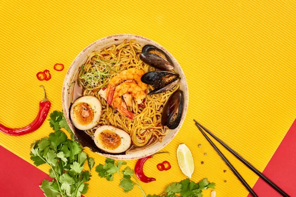 Vue de dessus des ramen de fruits de mer près des ingrédients frais et des baguettes sur la surface jaune et rouge — Photo de stock