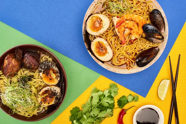 Vue de dessus des fruits de mer épicés et des ramen de viande près d'ingrédients frais, sauce soja et baguettes sur une surface multicolore — Photo de stock