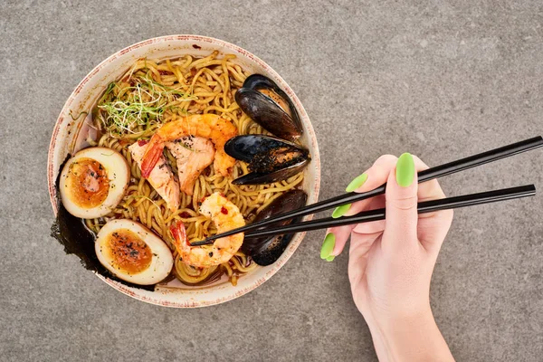 Cropped view of woman holding chopsticks near spicy seafood ramen on grey surface — Stock Photo