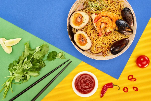 Vue de dessus des ramen épicés de fruits de mer près des ingrédients frais, sauce chili et baguettes sur la surface multicolore — Photo de stock