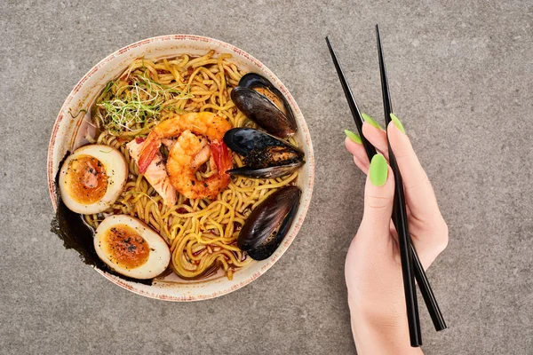 Cropped view of woman holding chopsticks near spicy seafood ramen on grey surface — Stock Photo
