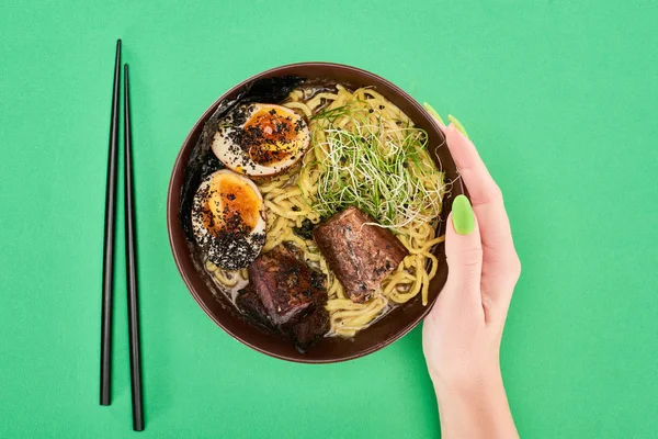 Cropped view of woman holding spicy meat ramen near chopsticks on green surface — Stock Photo