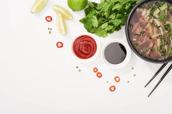 Top view of pho in bowl near chopsticks, lime, chili and soy sauces and coriander on white background — Stock Photo