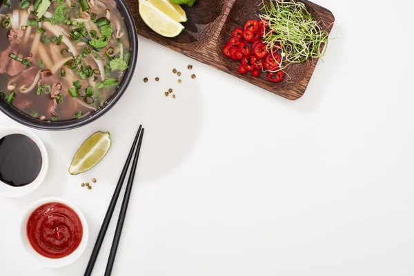 Top view of pho in bowl near chopsticks, lime, chili and soy sauces and coriander on white background — Stock Photo