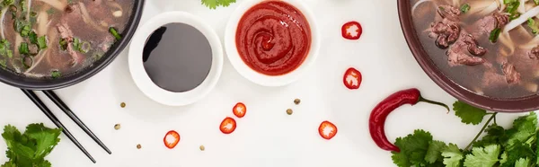 Top view of pho in bowls near chopsticks, chili and soy sauces and coriander on white background, panoramic shot — Stock Photo