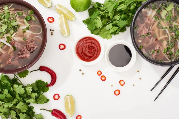 Top view of pho in bowls near chopsticks, lime, chili and soy sauces and coriander on white background — Stock Photo