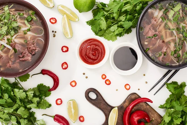 Top view of pho in bowls near chopsticks, lime, chili and soy sauces and coriander on white background — Stock Photo