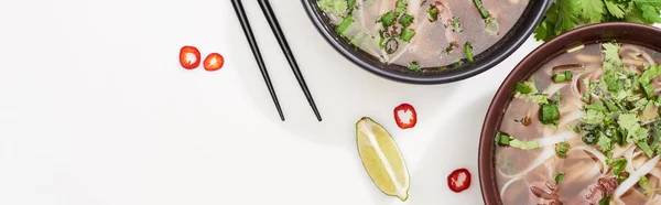 Top view of pho in bowl near chopsticks, lime, chili and soy sauces and coriander on white background, panoramic shot — Stock Photo