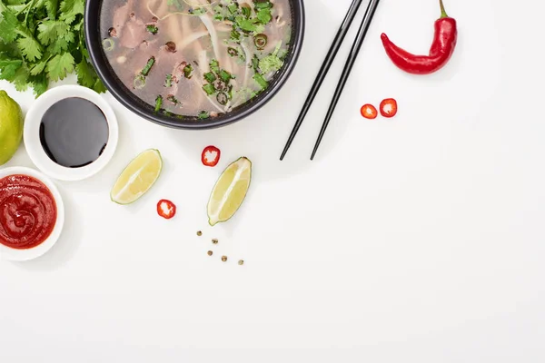 Top view of pho in bowl near chopsticks, lime, chili and soy sauces and coriander on white background — Stock Photo