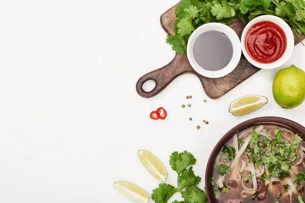 Верхний вид фо в миске рядом палочки для еды, лайм, чили и соевые соусы и кориандр на белом фоне — стоковое фото