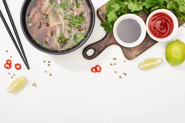 Top view of pho in bowl near chopsticks, lime, chili and soy sauces and coriander on white background — Stock Photo