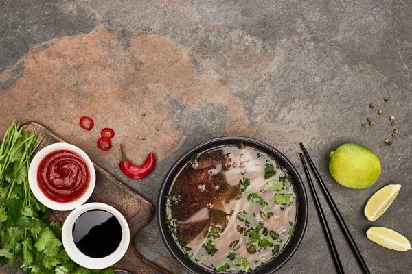 Vue du dessus du pho dans un bol près de baguettes, sauces citron vert, chili et soja et coriandre sur fond de pierre — Photo de stock