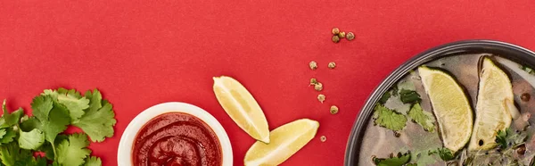 Top view of pho in bowl near lime, chili and soy sauces and coriander on red background, panoramic shot — Stock Photo