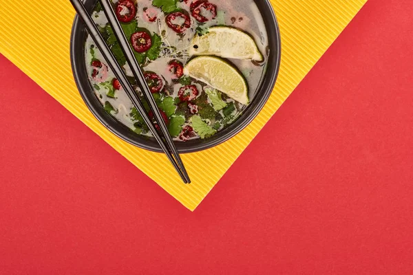 Верхний вид фо в миске с палочками для еды, лаймом, чили и кориандром на красном и желтом фоне — стоковое фото