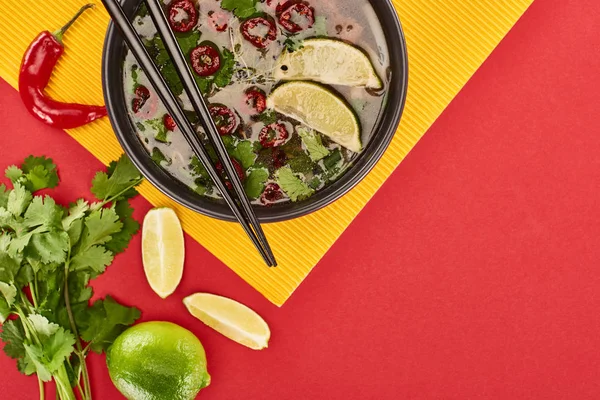 Top view of pho in bowl with chopsticks near lime, chili and coriander on red and yellow background — Stock Photo