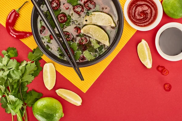 Vue du haut du pho dans un bol avec baguettes près des sauces, citron vert, chili et coriandre sur fond rouge et jaune — Photo de stock