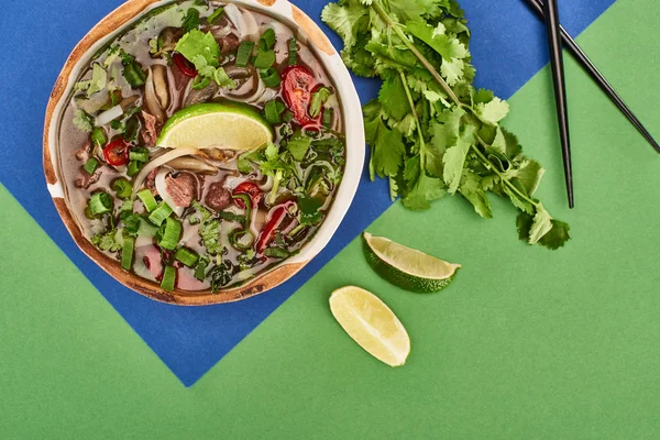 Верхний вид фо в миске рядом палочки для еды, лайм, кориандр на синем и зеленом фоне — стоковое фото