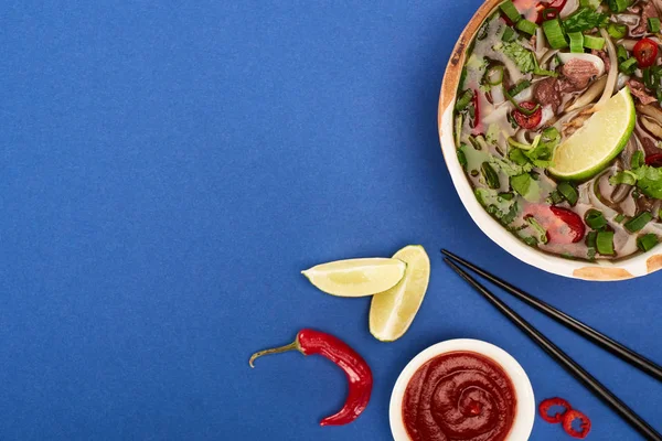 Vue du haut du pho dans un bol près des baguettes, citron vert, sauce chili sur fond bleu — Photo de stock