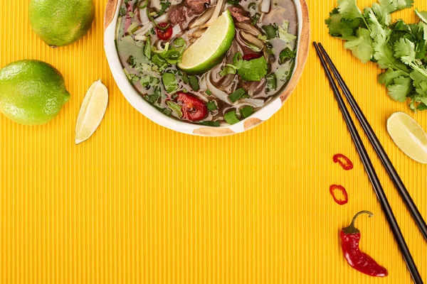 Vue du haut du pho dans un bol près des baguettes, de la lime, du chili et de la coriandre sur fond texturé jaune — Photo de stock