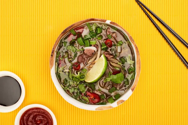 Top view of pho in bowl near chopsticks, chili and soy sauces on yellow textured background — Stock Photo