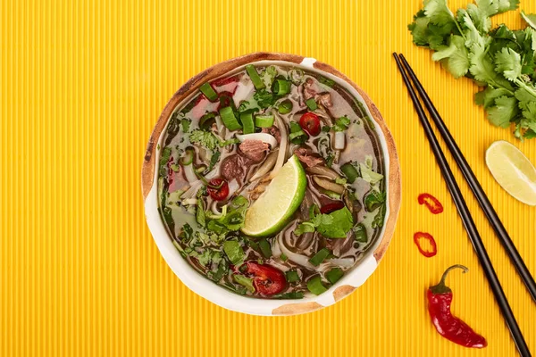 Top view of pho in bowl near chopsticks, lime, chili and coriander on yellow textured background — Stock Photo