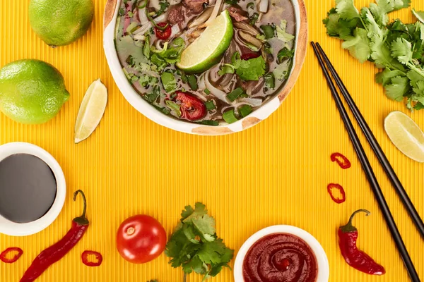 Top view of pho in bowl near chopsticks, lime, chili and soy sauces and coriander on yellow textured background — Stock Photo