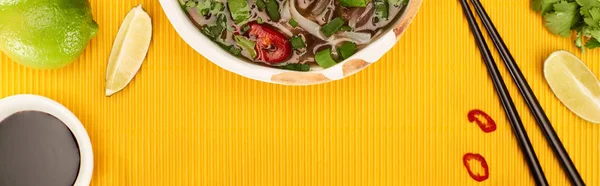 Top view of pho in bowl near chopsticks, lime, soy sauce and coriander on yellow textured background, panoramic shot — Stock Photo