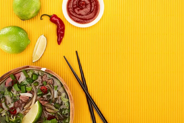 Vue du haut du pho dans un bol près des baguettes, citron vert, sauce chili sur fond texturé jaune — Photo de stock