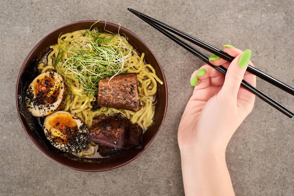 Vista recortada de la mujer sosteniendo palillos cerca de ramen con huevo y carne de res en un tazón sobre fondo gris - foto de stock