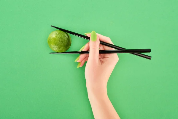 Cropped view of woman holding whole lime with chopsticks on green background — Stock Photo