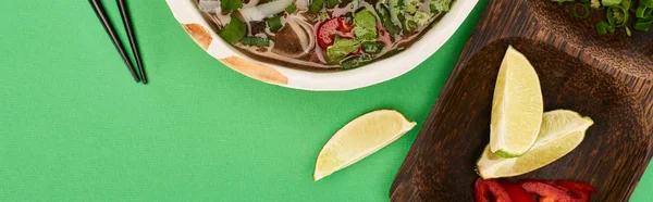 Top view of pho in bowl near chopsticks and wooden board with lime on green background, panoramic shot — Stock Photo