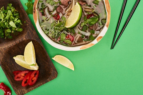 Top view of pho in bowl near chopsticks and wooden board with lime, chili and coriander on green background — Stock Photo