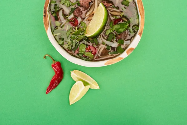 Top view of pho in bowl near lime, chili on green background — Stock Photo