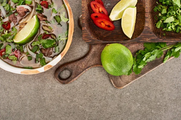 Top view of pho in bowl near lime, chili and coriander on wooden cutting board on grey background — Stock Photo