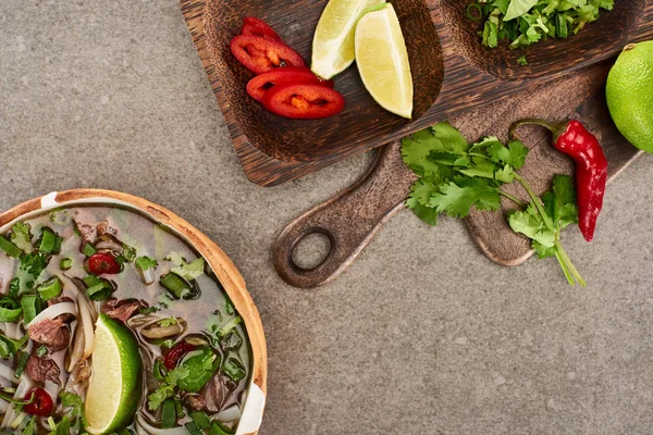 Top view of pho in bowl near lime, chili and coriander on wooden cutting board on grey background — Stock Photo