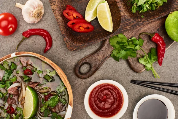 Top view of pho in bowl near ingredients, chili and soy sauces and chopsticks on grey background — Stock Photo