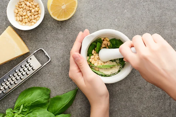 Vue recadrée de la femme broyage des ingrédients crus de sauce pesto en livre sur la surface grise — Photo de stock