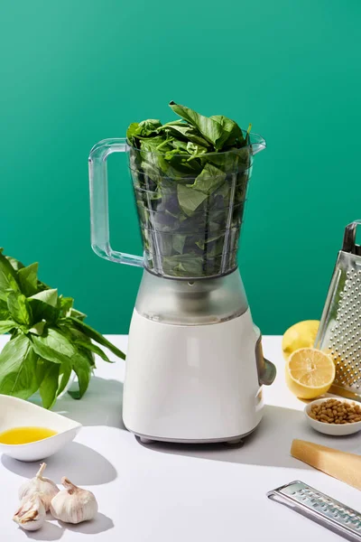 Sauce pesto ingrédients crus et robot culinaire avec feuilles de basilic sur table blanche isolé sur vert — Photo de stock