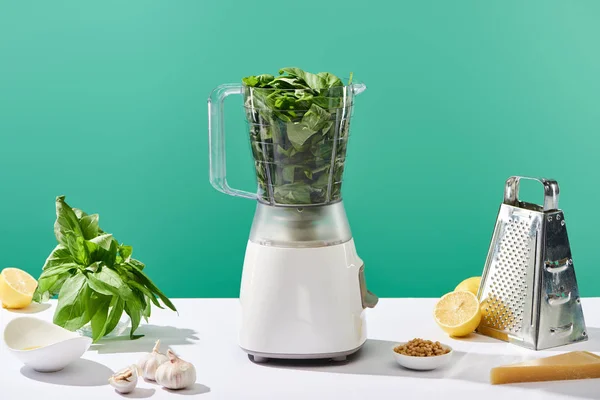 Molho pesto ingredientes crus e processador de alimentos com folhas de manjericão na mesa branca isolada em verde — Fotografia de Stock