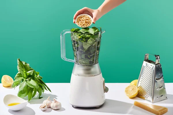 Cropped view of woman adding pine nuts to basil leaves in food processor near pesto sauce raw ingredients on white table isolated on green — Stock Photo