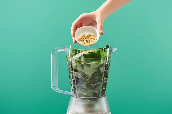 Cropped view of woman adding pine nuts to basil leaves in food processor isolated on green — Stock Photo