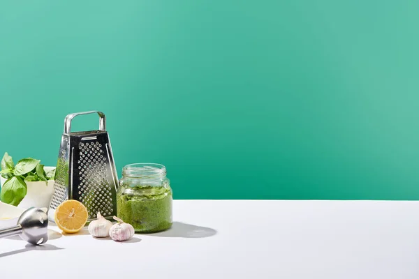 Salsa di pesto in vasetto vicino agli ingredienti, frullatore e grattugia su tavolo bianco isolato su verde — Foto stock