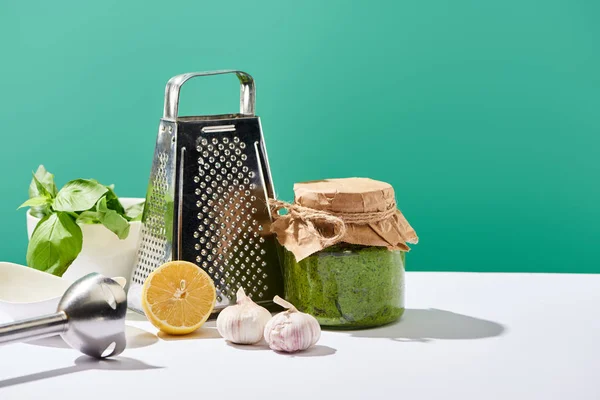 Sauce pesto dans un bocal près des ingrédients, mélangeur et râpe sur table blanche isolée sur vert — Photo de stock