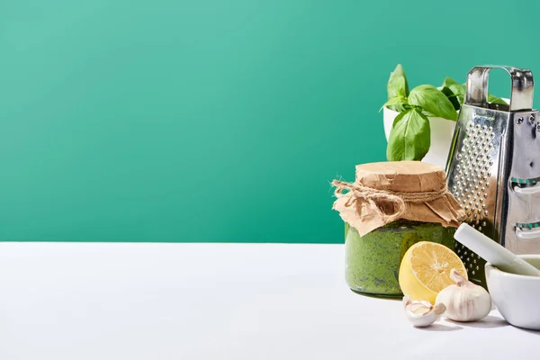 Соус песто в банці біля інгредієнтів і тертя на білому столі ізольовані на зеленому — стокове фото