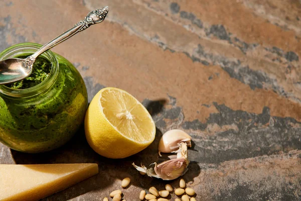 Molho pesto em frasco de vidro com colher perto de ingredientes na superfície de pedra — Fotografia de Stock