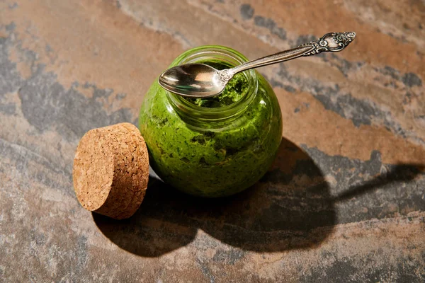 Salsa pesto en frasco de vidrio con cuchara cerca de corcho en la superficie de piedra - foto de stock