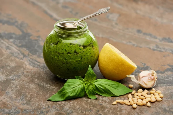 Pesto sauce in glass jar with spoon near ingredients on stone surface — Stock Photo