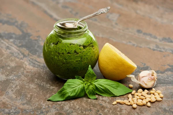 Molho pesto em frasco de vidro com colher perto de ingredientes na superfície de pedra — Fotografia de Stock