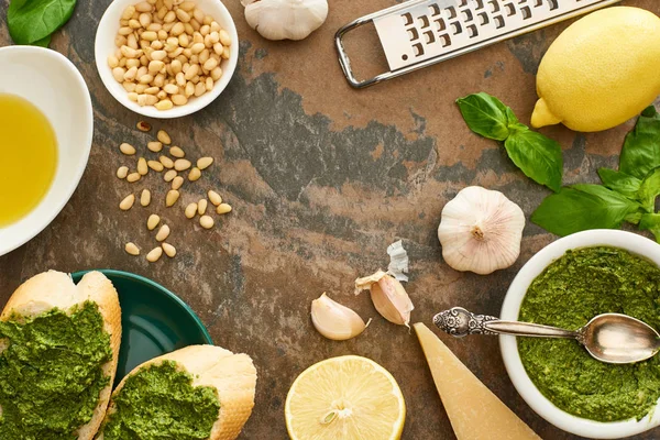 Vista superior de fatias de baguete com molho de pesto na placa perto de ingredientes frescos e utensílios de cozinha na superfície de pedra — Fotografia de Stock